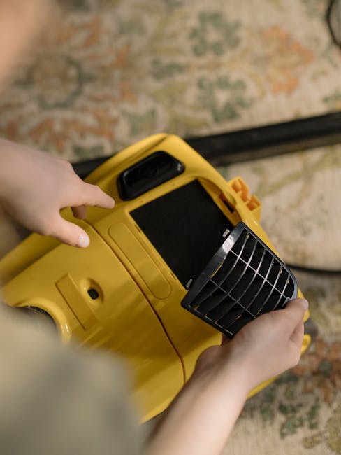 A person operating a yellow portable carpet cleaning machine with a black handle and a vented front panel, positioned on a patterned rug with floral and geometric design in shades of beige, green, and