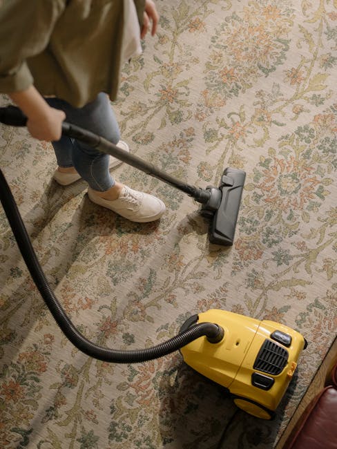 A person operating a yellow portable carpet cleaning machine with a black handle and a vented front panel, positioned on a patterned rug with floral and geometric design in shades of beige, green, and